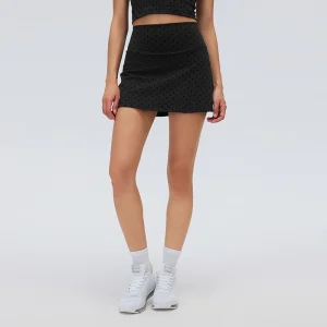 Front view of a model wearing a black flocked polka dot tennis skort with a wide high-rise waistband and A-line mini skirt.